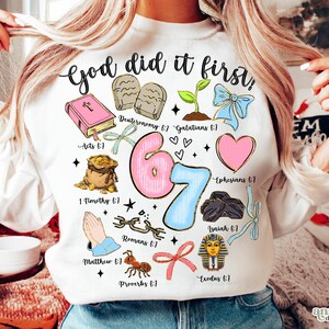 Könnte beinhalten: Weißes Sweatshirt mit dem Text "God did it first" und farbenfrohen Illustrationen, darunter ein rosa Buch, ein rosa Herz und eine blaue Schleife. Die Zahl "67" ist in Rosa und Blau.