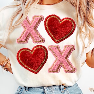 Op de afbeelding: Cr&egrave;mekleurige sweatshirt met roze en gouden letters X en rode harten met gouden rand. Het ontwerp is een speelse opstelling van de letters X en O, waardoor een Valentijnsdagthema ontstaat. De sweatshirt is een casual, comfortabele stijl.