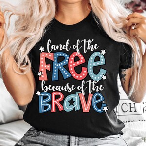 以下が含まれることがあります： 白い星が散りばめられた、赤、白、青の文字で「land of the free because of the brave」と書かれた黒いTシャツ。