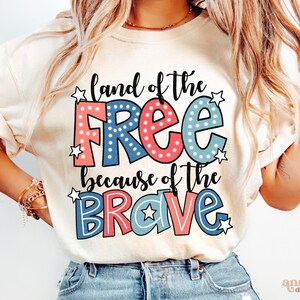 以下が含まれることがあります： 「land of the free because of the brave」と書かれた赤、白、青のグラフィックがプリントされた白いTシャツ。グラフィックは、星と水玉模様の漫画風です。
