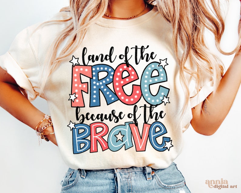 以下が含まれることがあります： 「land of the free because of the brave」と書かれた赤、白、青のグラフィックがプリントされた白いTシャツ。グラフィックは、星と水玉模様の漫画風です。