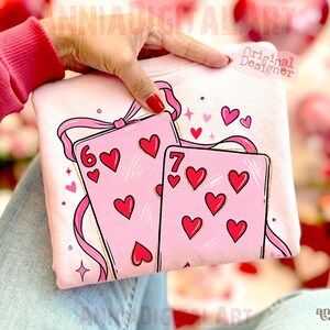 May include: A light pink sweatshirt featuring a design of two playing cards with red hearts. The cards are decorated with pink ribbons and hearts. The number six and seven of hearts are visible on the cards. The words "Original Designer" are visible.