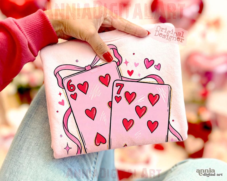 May include: A light pink sweatshirt featuring a design of two playing cards with red hearts. The cards are decorated with pink ribbons and hearts. The number six and seven of hearts are visible on the cards. The words "Original Designer" are visible.