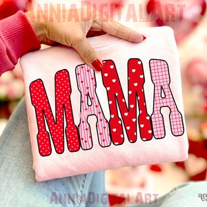 May include: A light pink sweatshirt with the word "MAMA" in red and pink letters. The letters are decorated with polka dots, hearts, and a grid pattern. The sweatshirt is held by a person.