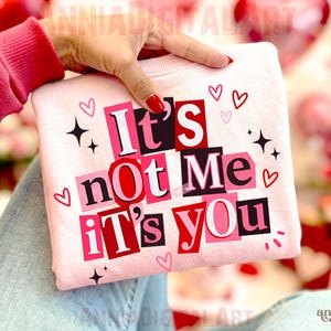 Puede incluir: Una sudadera rosa claro con el texto "It's not Me It's You" en un diseño estilo collage. El diseño incluye cuadrados rosas y rojos, corazones y estrellas. La sudadera es sostenida por una persona que lleva un suéter rojo.