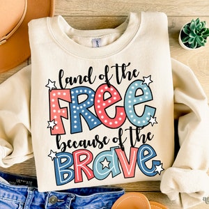 以下が含まれることがあります： クリーム色のスウェットシャツに、「land of the free because of the brave」という文字が赤、青、白で書かれており、白い星が散りばめられています。文字は太字で、漫画のようなフォントです。