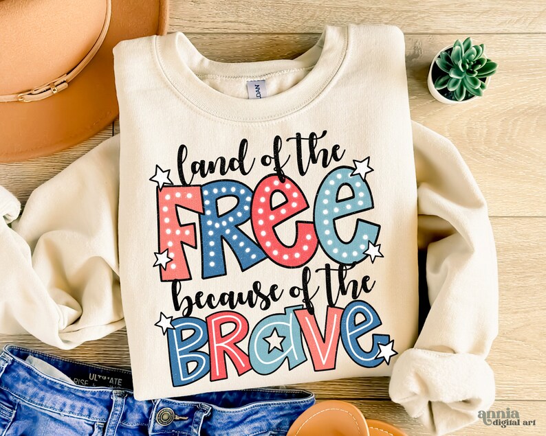 以下が含まれることがあります： クリーム色のスウェットシャツに、「land of the free because of the brave」という文字が赤、青、白で書かれており、白い星が散りばめられています。文字は太字で、漫画のようなフォントです。