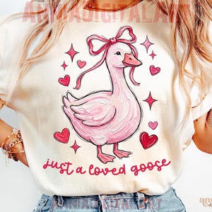 Pu&ograve; includere: T-shirt color crema con un'illustrazione di un'oca rosa con un fiocco, circondata da cuori e stelle. Il testo "just a loved goose" &egrave; scritto in corsivo. Un design carino e stravagante.