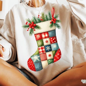 May include: A cream-colored sweatshirt featuring a colorful Christmas stocking design. The stocking is decorated with patchwork squares in red, green, blue, and white, adorned with snowflakes, stars, and polka dots. A sprig of greenery and red berries tops the stocking.