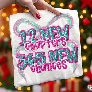 May include: White t-shirt with the text "12 NEW chapters 365 NEW chances" in teal and pink glitter. The design includes a teal and pink ribbon-like graphic. The shirt is held up by a hand with red nail polish.
