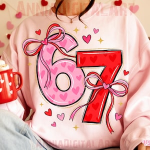 May include: Pink sweatshirt with a large graphic of the numbers "67" in pink and red, decorated with hearts and bows. The number "6" is pink with heart patterns, and the number "7" is red with heart patterns. Small hearts and stars are scattered around the numbers.