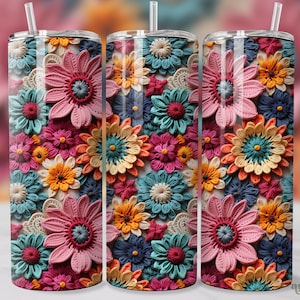 May include: A set of three stainless steel tumblers featuring a colourful floral pattern. The pattern includes a variety of flowers in shades of pink, blue, yellow, and orange. The flowers are made from yarn or string and have a textured appearance.