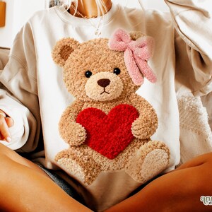 May include: Cream-colored sweatshirt featuring a plush teddy bear design. The bear holds a red heart and has a pink bow. The sweatshirt is a cozy, casual style, perfect for a relaxed look.