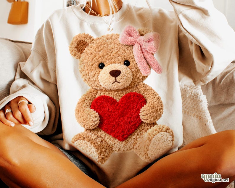 May include: Cream-colored sweatshirt featuring a plush teddy bear design. The bear holds a red heart and has a pink bow. The sweatshirt is a cozy, casual style, perfect for a relaxed look.