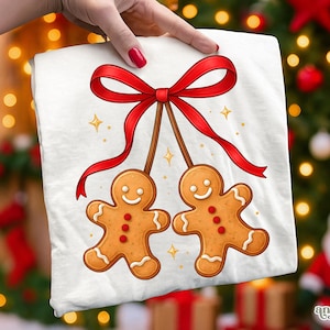 Puede incluir: Camiseta blanca con un diseño festivo de dos hombres de jengibre colgando de un lazo rojo. Los hombres de jengibre tienen caras sonrientes y están decorados con botones rojos. Estrellas doradas salpican el diseño.