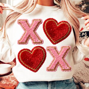 Op de afbeelding: Witte sweatshirt met roze en gouden "X" en rode hart patches. De patches hebben een fuzzy textuur en zijn goud omrand. Het ontwerp is gecentreerd op de voorkant van het shirt.