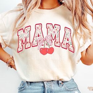 May include: A white t-shirt with the word "MAMA" printed in red with a floral pattern. The word is outlined in red and has a pink bow and two red cherries below it.