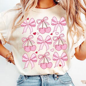 May include: A white t-shirt with a pink and red cherry and bow design. The design features multiple cherries with stems and leaves, each tied with a pink bow. Small red hearts are scattered throughout the design.