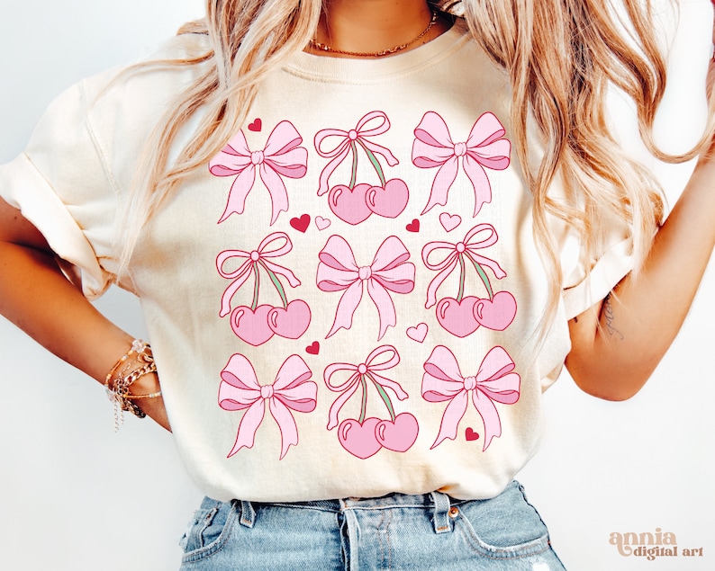 May include: A white t-shirt with a pink and red cherry and bow design. The design features multiple cherries with stems and leaves, each tied with a pink bow. Small red hearts are scattered throughout the design.