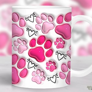 May include: White ceramic mug with a pink paw print design. The paw prints are in a 3D style and have a glossy finish. The mug has a black handle and a white background.