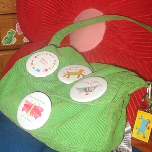 Può includere: Una borsa a tracolla in velluto a coste verde brillante con quattro spille rotonde decorate con varie illustrazioni e testi, tra cui una farfalla rosa, un cane giallo, un piccione e lettere colorate, oltre a un portachiavi rettangolare giallo.