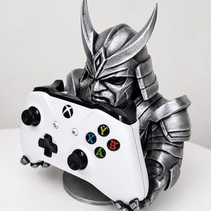 May include: A silver samurai bust holding a white Xbox controller. The controller has black accents and coloured buttons. The bust has detailed armour and a stern expression, with a round base.