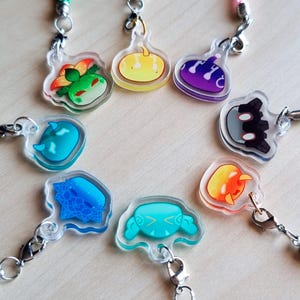 Puede incluir: Una colección de colgantes de acrílico transparente con diseños de dibujos animados coloridos, cada uno unido a un cordón. Los colgantes presentan varias formas y personajes en tonos de verde, amarillo, morado, azul, naranja y negro. Los cordones son de varios colores.