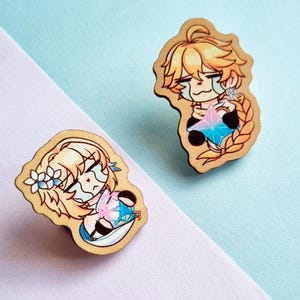 Pode incluir: Dois pinos de madeira com personagens em estilo anime com cabelo loiro e expressões de choro. Um pino mostra um personagem segurando uma gema azul e rosa. O outro pino mostra um personagem com flores no cabelo. Fundo azul claro e branco.