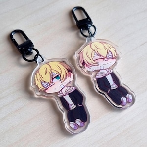 Op de afbeelding: Twee transparante acryl sleutelhangers met een stripfiguur met blond haar, verbanden en een zwarte outfit. Elke sleutelhanger heeft een zwarte sluiting. Het personage heeft een neutrale uitdrukking en bevindt zich in een ontspannen pose.