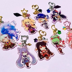 May include: A selection of acrylic keyrings with vibrant anime-style character designs. Each keyring showcases a distinct character illustration, with gold-tone hardware and decorative charms. The keyrings are displayed on a white surface.