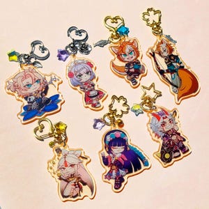 Pode incluir: Uma coleção de sete chaveiros de acrílico com ilustrações coloridas de personagens em estilo anime. Cada pingente inclui um fecho de metal em formato de coração ou estrela, decorado com pequenas contas e berloques presos aos anéis.
