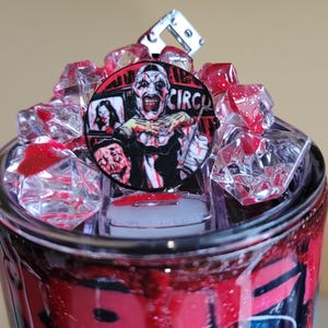 May include: A tumbler with a horror theme, featuring a clown design. The top is adorned with red, crystal-like accents and a circular emblem with the word "CIRCUS". The tumbler has a black and red design.