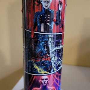 May include: A stainless steel tumbler with a red, black, and blue design featuring characters from the horror movie "Hellraiser". The tumbler has a clear lid and a black straw.