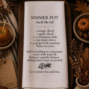 May include: A white rectangular kitchen towel featuring black printed text that reads SIMMER POT Smells like Fall, followed by a recipe list of ingredients including orange, apple, cinnamon sticks, cloves, rosemary, and water, with instructions.