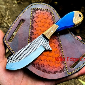 May include: A blue and wood handled knife with a serrated blade and a leather sheath. The sheath has a woven pattern and the text "Wild Edge Crafts" is visible.