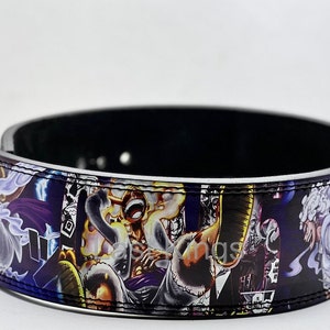 May include: A black leather weightlifting belt with a colourful anime print featuring characters from the popular manga series One Piece. The belt has a silver buckle and white stitching.