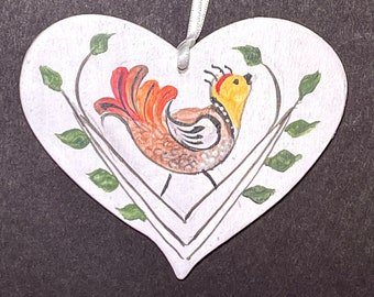 May include: White heart-shaped ornament with a painted bird with orange, yellow, and brown feathers. The bird is perched on a branch with green leaves.