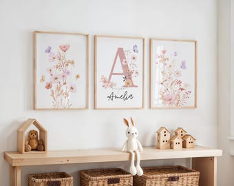 May include: Three framed floral art prints with a light wood frame. One print features the letter "A" with the name "Amelia" below. The other two prints have floral and butterfly designs. A wooden shelf below holds decorative items.