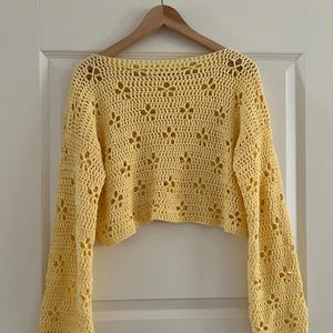 May include: A yellow crochet top with long sleeves and a floral pattern. The top is hanging on a wooden hanger against a white door. The crochet design features openwork details.
