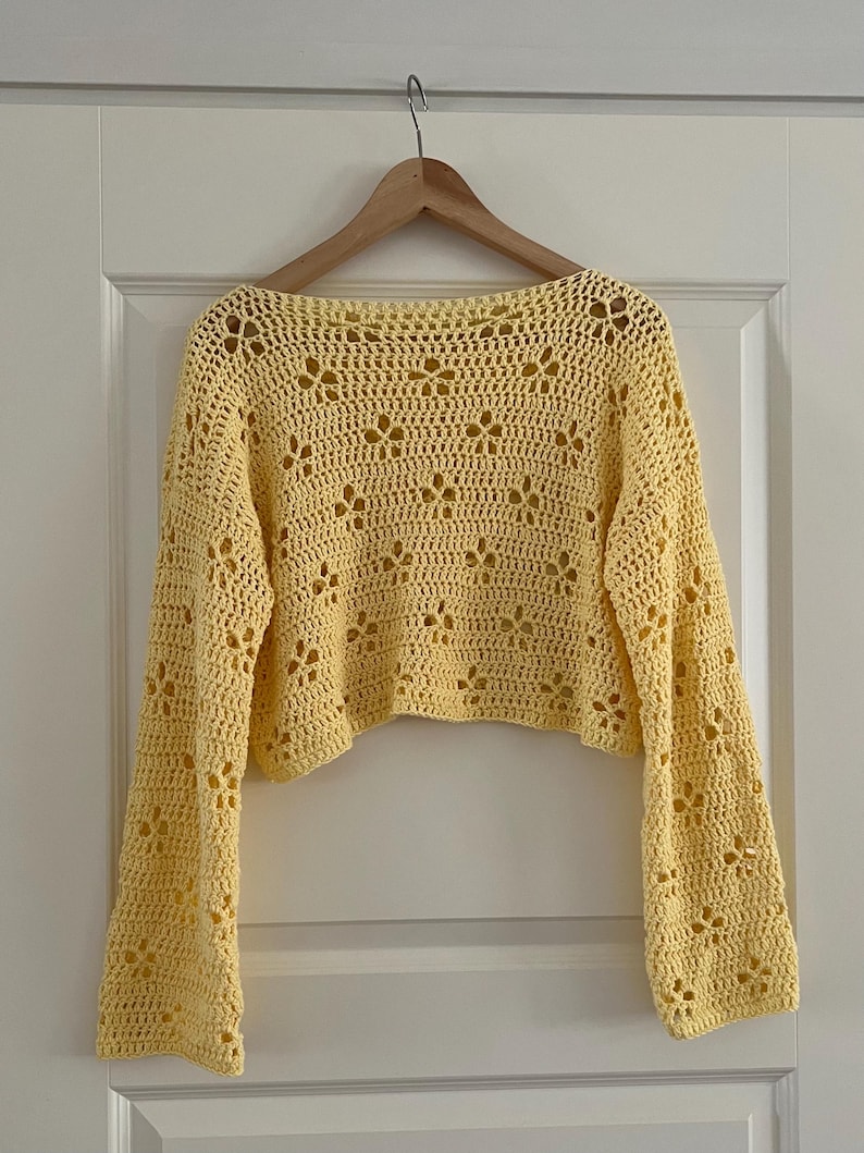 May include: A yellow crochet top with long sleeves and a floral pattern. The top is hanging on a wooden hanger against a white door. The crochet design features openwork details.