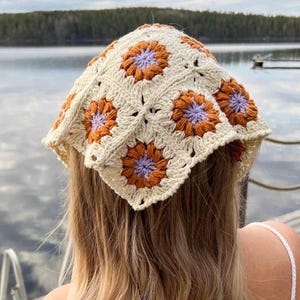 May include: Cream-colored crochet headscarf featuring a floral pattern with orange and purple accents. The square scarf is made of yarn and is worn on the head. The head covering is a handmade accessory.
