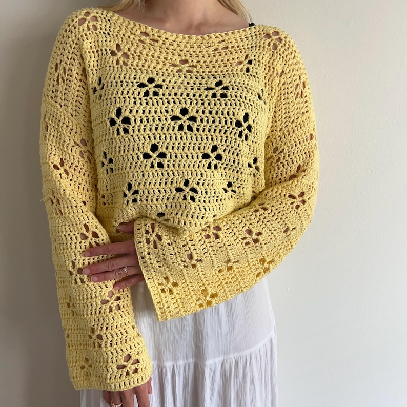 May include: A light yellow, long-sleeved crochet top with a floral pattern. The top has a boat neck and flared sleeves. The crochet design features openwork details, creating a delicate and airy look. The top is paired with a white skirt.