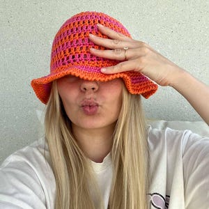 Puede incluir: Sombrero de pescador de ganchillo hecho a mano en tonos vibrantes de rosa y naranja. El sombrero presenta un ala ancha, ligeramente rizada y un diseño de tejido abierto. Un accesorio de moda para protegerse del sol.