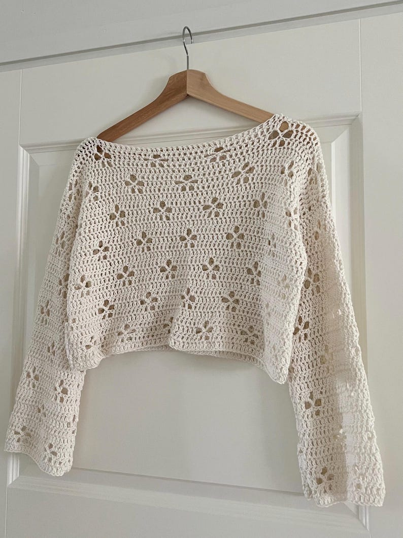 May include: Cream-colored, long-sleeved crochet top with a floral pattern. The top is hanging on a wooden hanger against a white door. The crochet design features openwork details, creating a delicate and airy look.