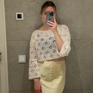 May include: Cream-colored, open-weave crochet top with bell sleeves. The top features a floral pattern and a cropped design. Paired with a light yellow satin skirt. The image is taken in front of a gray tiled wall.