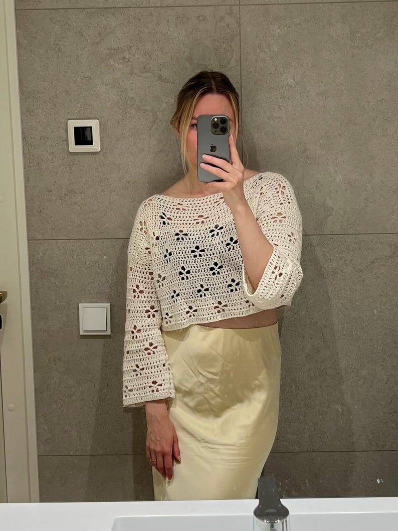 May include: Cream-colored, open-weave crochet top with bell sleeves. The top features a floral pattern and a cropped design. Paired with a light yellow satin skirt. The image is taken in front of a gray tiled wall.