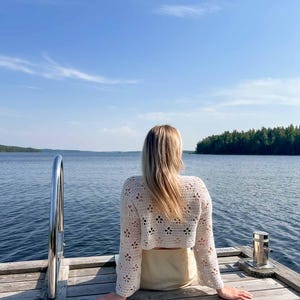 May include: A cream-colored, open-weave crochet top with long sleeves. The top features a floral pattern and is paired with a light-colored skirt. The person is sitting on a wooden dock overlooking a lake under a blue sky.
