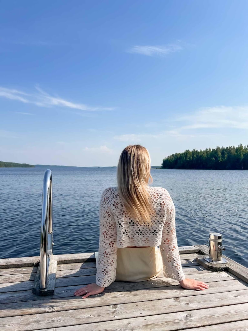 May include: A cream-colored, open-weave crochet top with long sleeves. The top features a floral pattern and is paired with a light-colored skirt. The person is sitting on a wooden dock overlooking a lake under a blue sky.