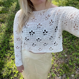 May include: Cream-colored, open-weave crochet top with long sleeves. The top features a floral pattern and a cropped design. The model is wearing a cream-colored skirt. The background is green with natural light.