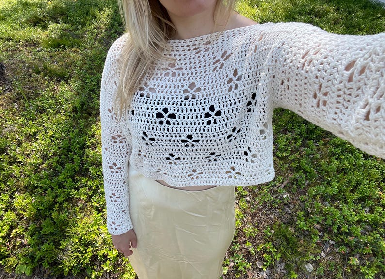 May include: Cream-colored, open-weave crochet top with long sleeves. The top features a floral pattern and a cropped design. The model is wearing a cream-colored skirt. The background is green with natural light.